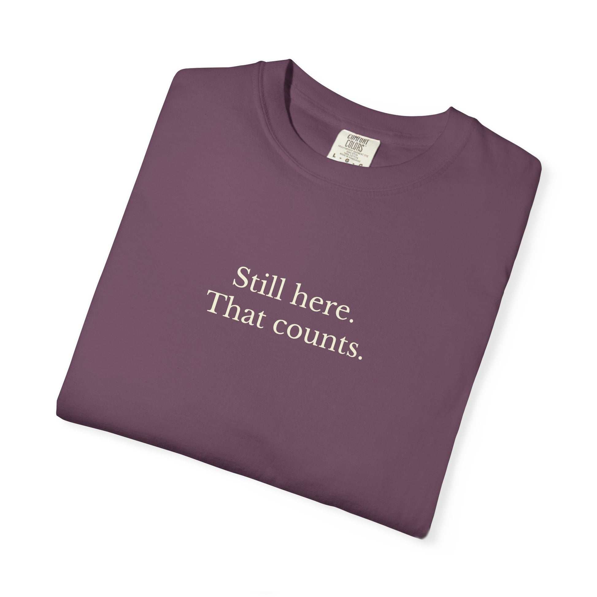 Folded purple t-shirt with text 'Still here. That counts.' on a white background