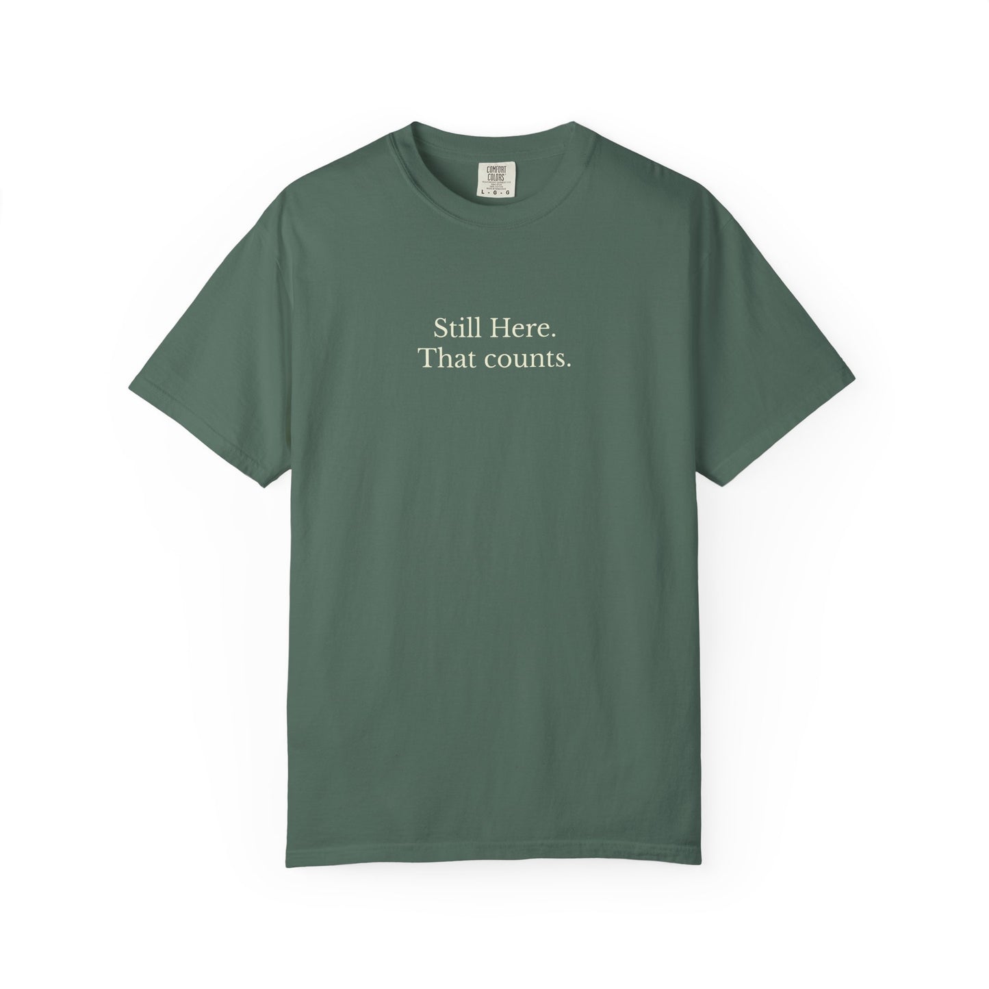 Green t-shirt with text 'Still Here. That counts.' on a white background