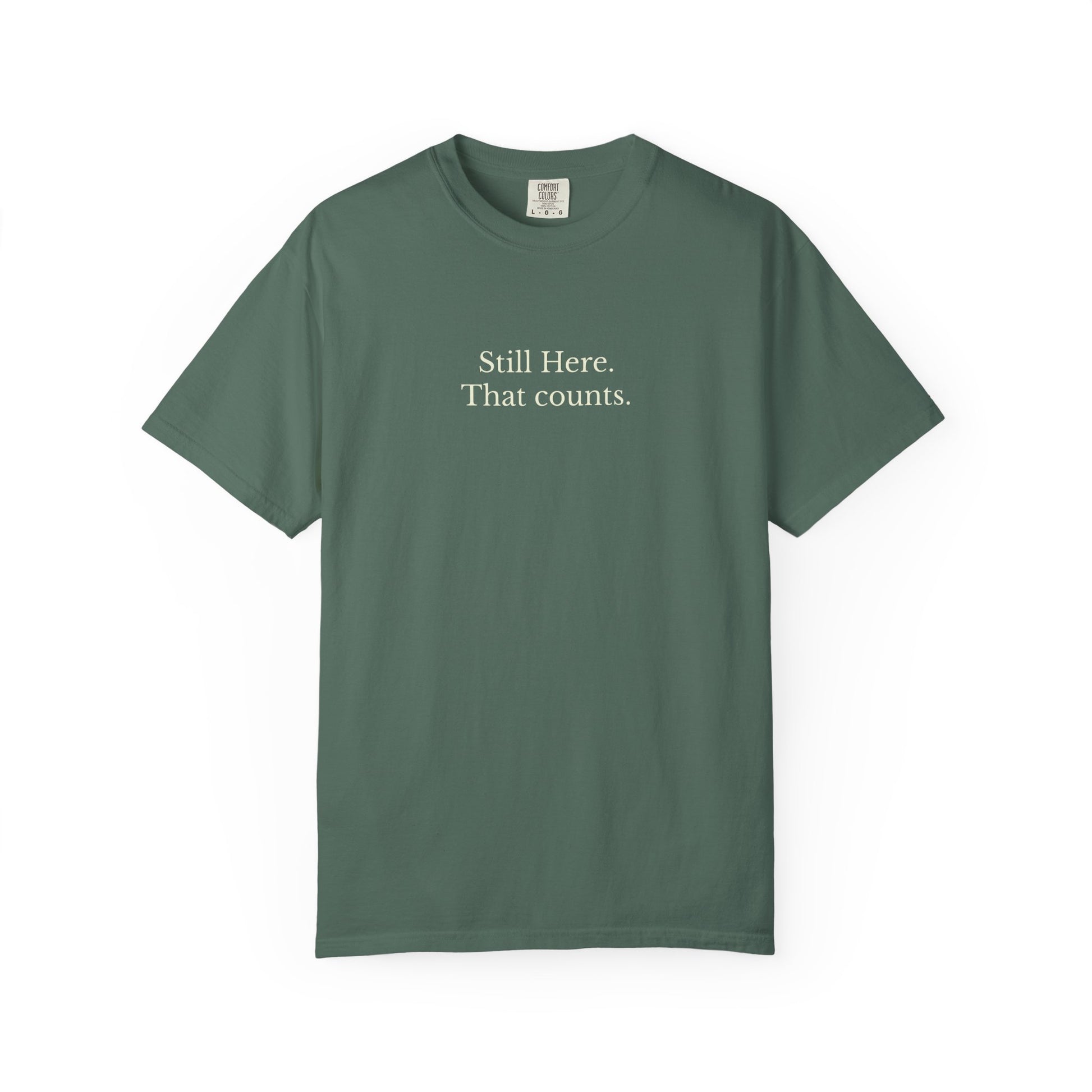 Green t-shirt with text 'Still Here. That counts.' on a white background