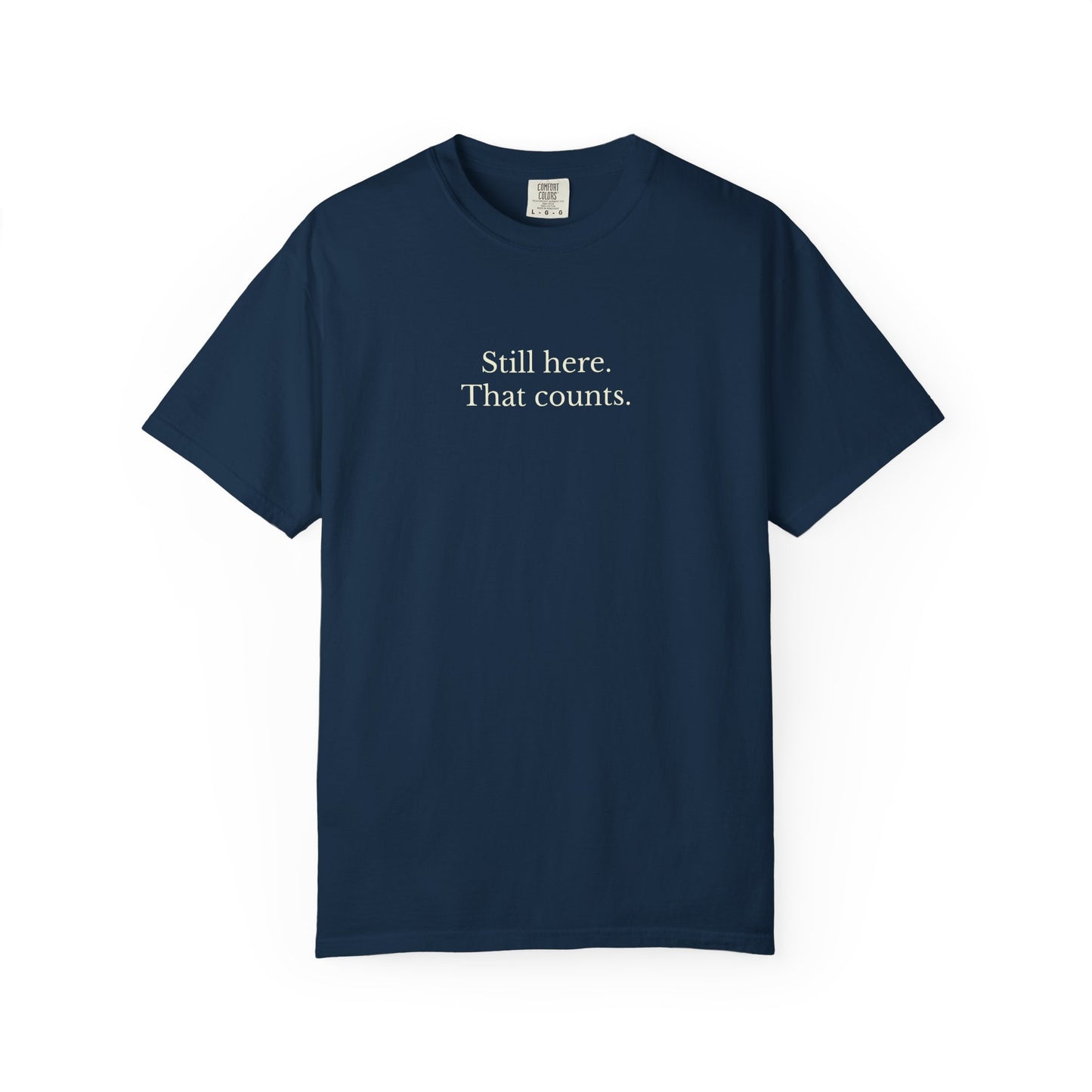 Navy blue t-shirt with white text 'Still here. That counts.' on a white background