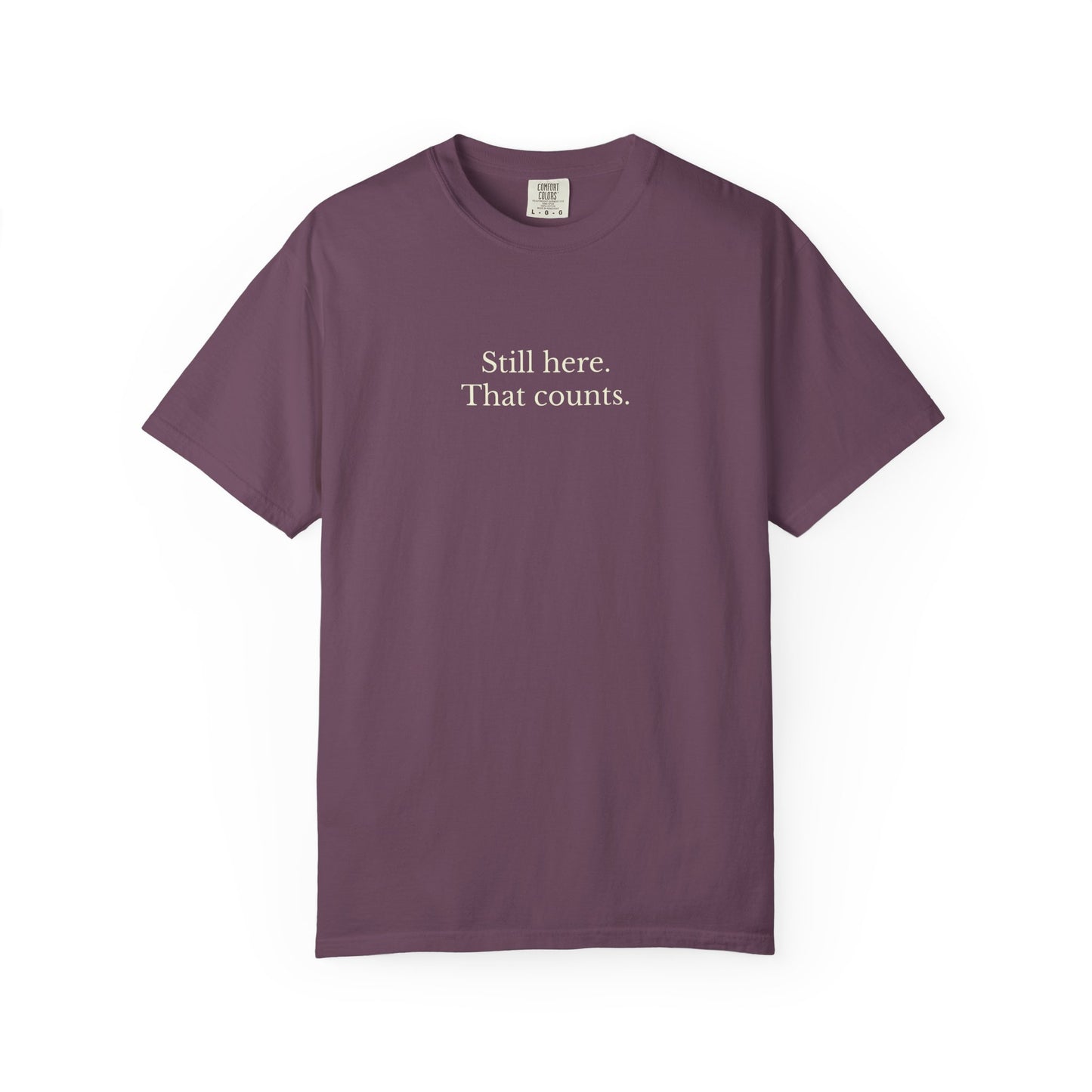 Purple t-shirt with text 'Still here. That counts.' on a white background
