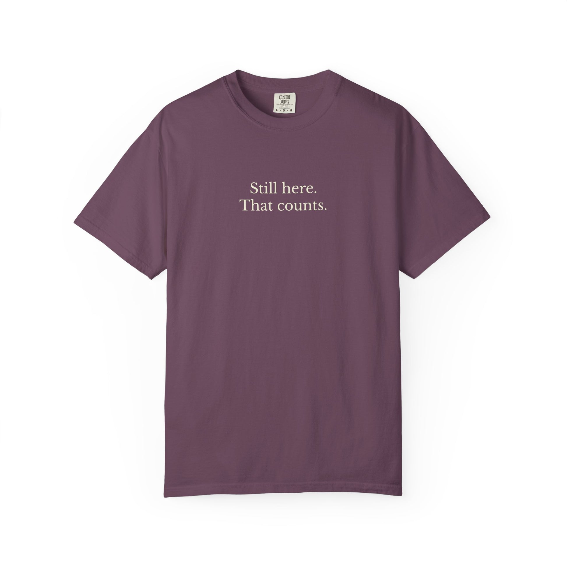 Purple t-shirt with text 'Still here. That counts.' on a white background