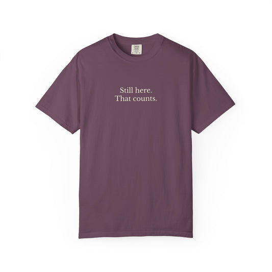 Purple t-shirt with text 'Still here. That counts.' on a white background