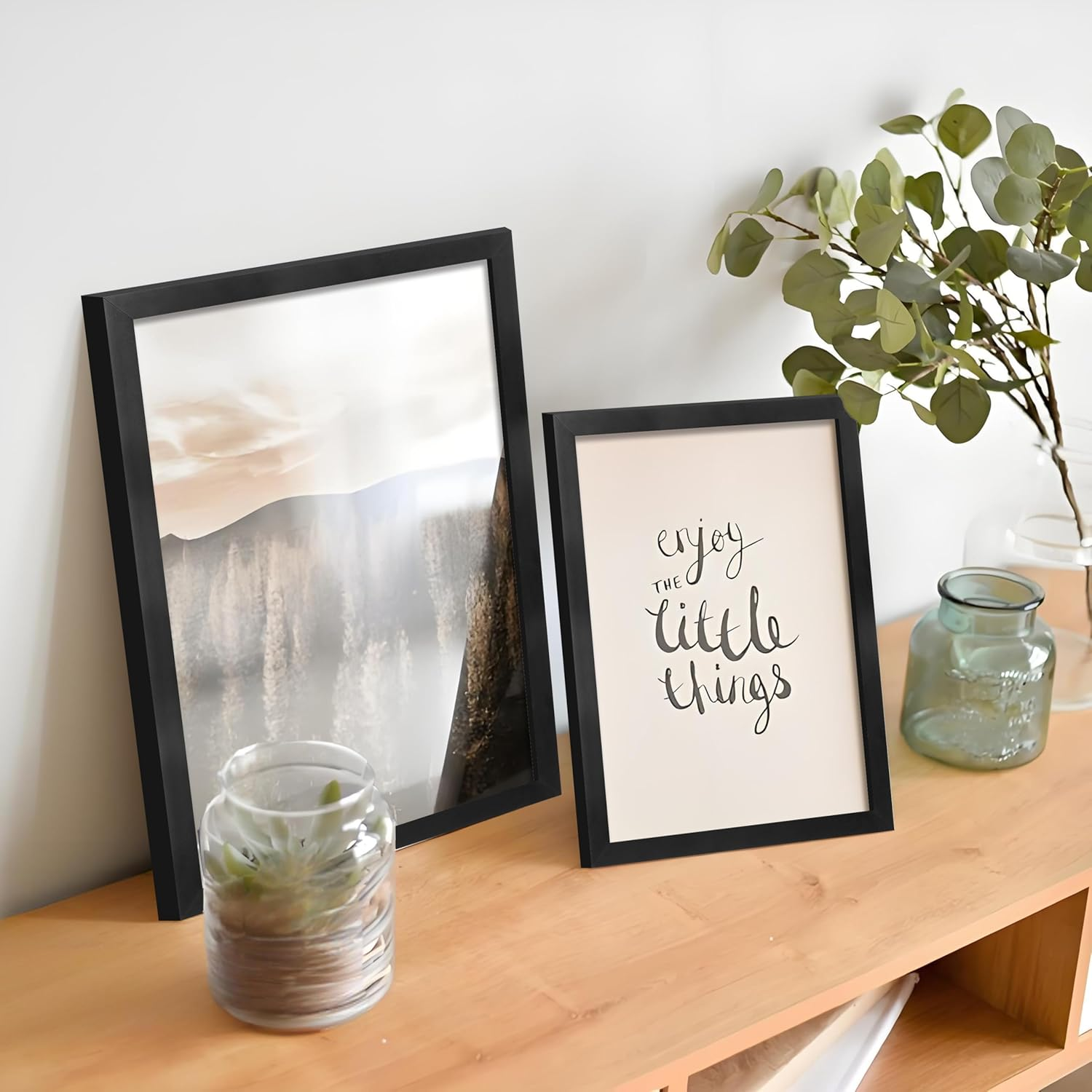 Two framed prints on a wooden surface with decorative jars and a plant.