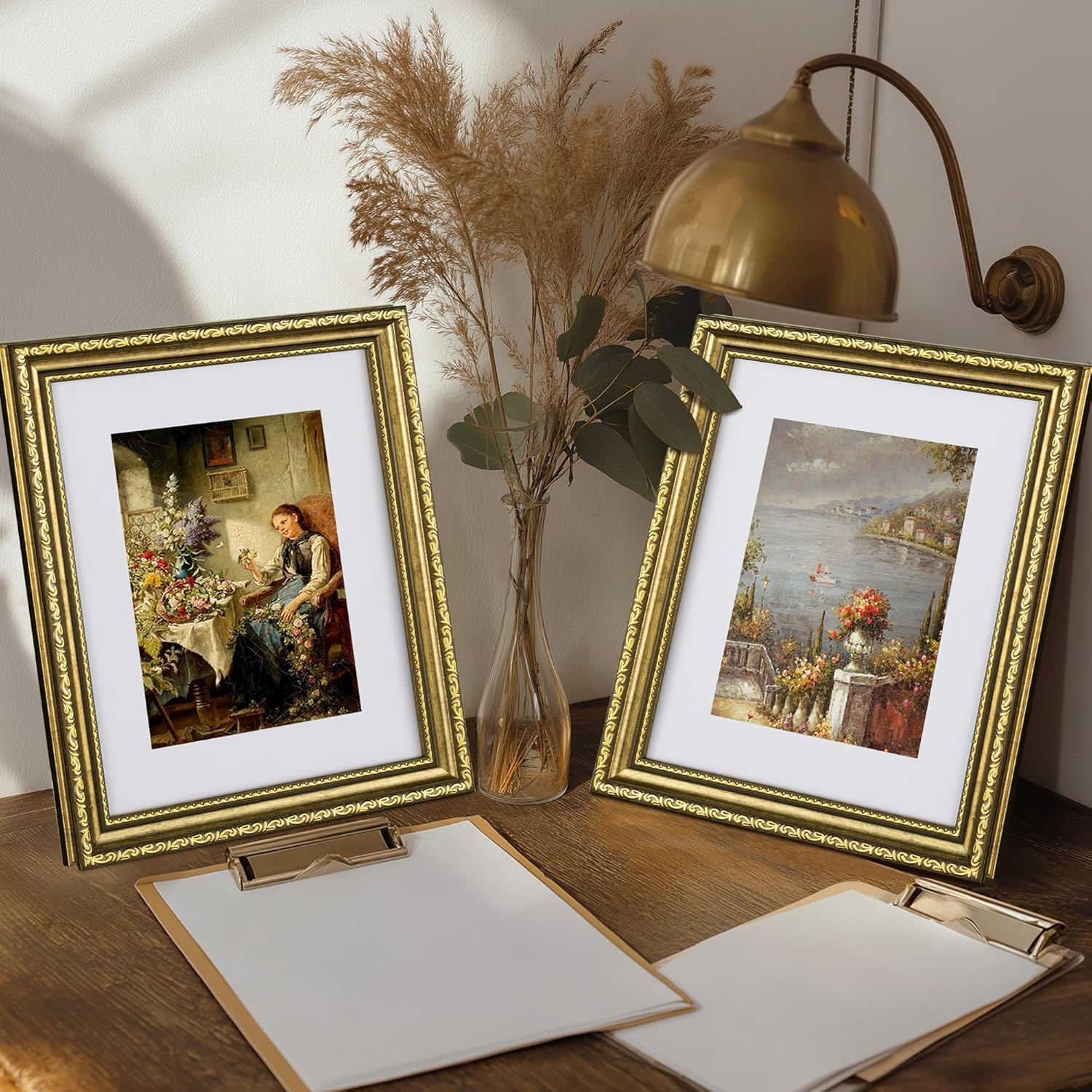 Two framed artworks on a wooden surface with a vase of dried plants and a lamp in the background.