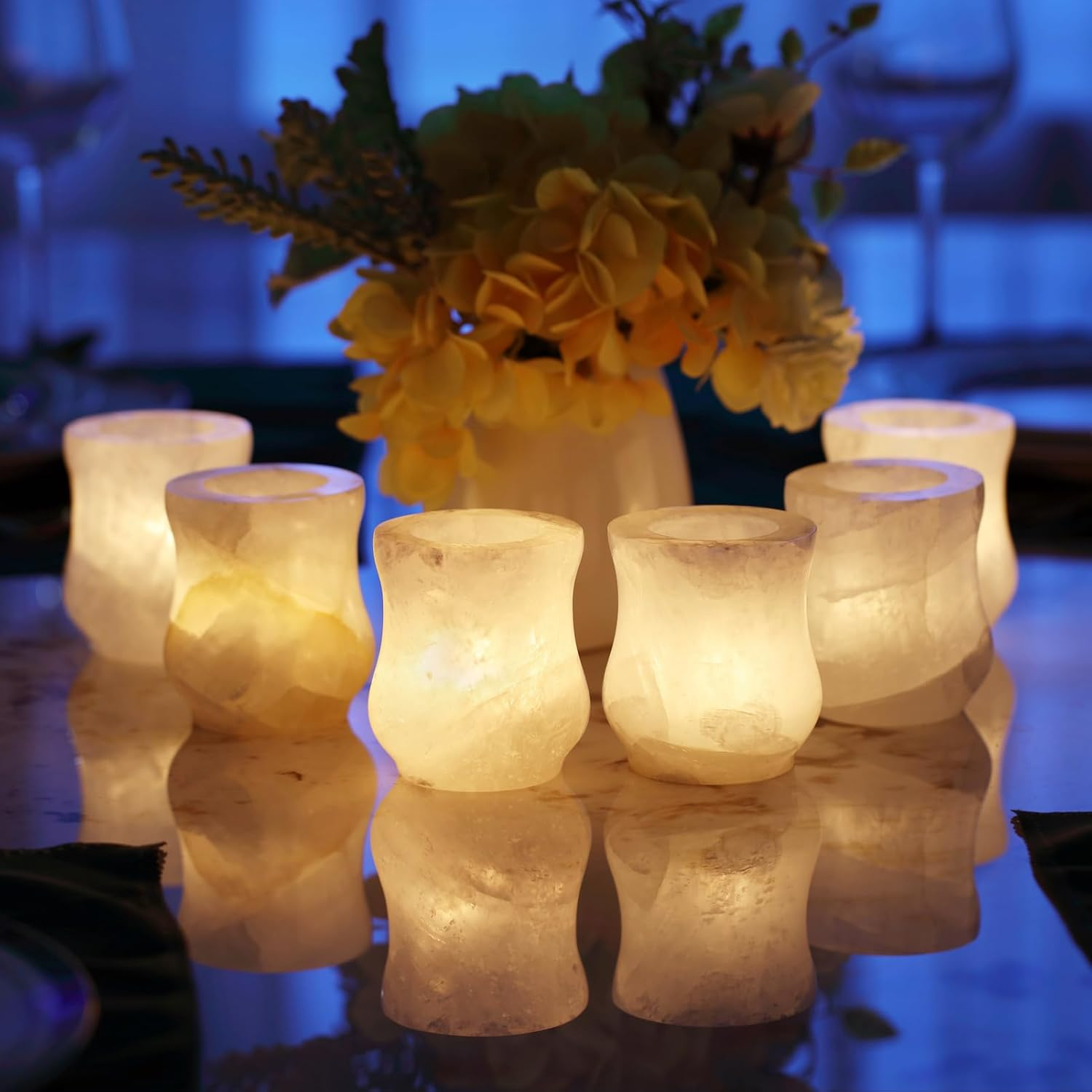 Set of lit candle holders with a vase of yellow flowers on a reflective surface.